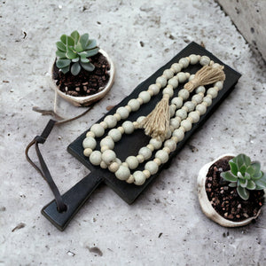 White beaded necklace with tassel on a black stand, surrounded by small potted plants on a textured surface.