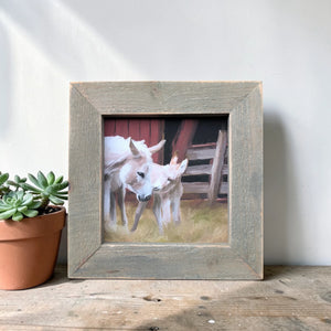 Framed picture of two horses on a wooden surface with a plant next to it.