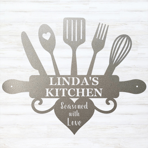 A silver personalized kitchen sign featuring silhouettes of kitchen utensils (knife, spoon with heart, spatula, fork, whisk, and rolling pin) arranged around the text 'LINDA'S KITCHEN' on a rolling pin, with a heart below reading 'Seasoned with Love'. The sign is a dark metallic gray color and is displayed against a white wood-plank background.
