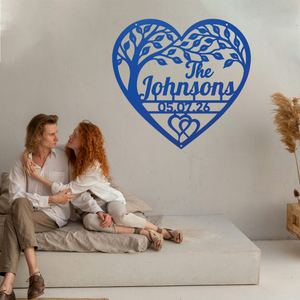 A couple sits on a low concrete bench against a white wall, with a large blue heart-shaped tree cutout wall decal behind them featuring the personalized text 'The Johnsons' and the date '05.07.