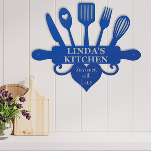 A blue metal kitchen wall sign shaped like kitchen utensils with the text 'LINDA'S KITCHEN' and 'Seasoned with Love' in a heart, mounted on a white paneled wall above a shelf with wooden cutting boards and flowers.