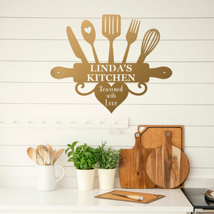 A decorative wall sign shaped like kitchen utensils and a rolling pin with the text 'LINDA'S KITCHEN' and 'Seasoned with Love' displayed above a kitchen counter with wooden cutting boards, plants, and utensils.