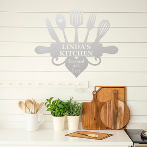 A gray kitchen-themed wall decal reading 'LINDA'S KITCHEN' with utensil silhouettes and the phrase 'Seasoned with Love,' displayed above a white countertop with wooden cutting boards, plants, and utensils in a bright kitchen setting.