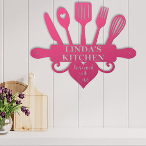 A pink metal kitchen wall sign shaped like a rolling pin with kitchen utensils and a heart, mounted on a white paneled wall above a countertop. The sign reads 'LINDA'S KITCHEN' and 'Seasoned with Love'.