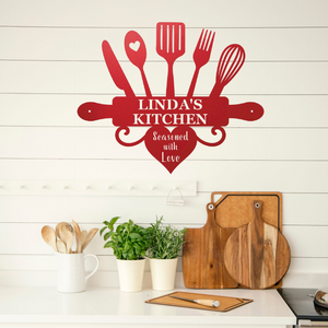 A red metal kitchen wall sign shaped like a rolling pin with utensils and the words 'LINDA'S KITCHEN' and 'Seasoned with Love' displayed on a white shiplap wall above a kitchen counter with cutting boards and plants.