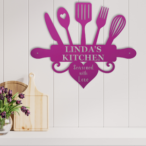 A personalized metal kitchen wall sign in bright magenta, featuring kitchen utensils and a rolling pin with the text 'LINDA'S KITCHEN' and 'Seasoned with Love'. The sign is displayed on a white paneled wall above a kitchen counter with wooden cutting boards and a vase of purple tulips.