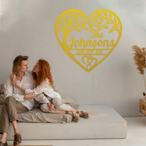 Two people sitting on a low concrete bench against a white wall, with a large personalized yellow heart-shaped tree wall sign behind them with a personalized name and date.