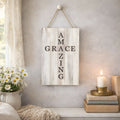 Decorative sign with 'AMAZING GRACE ZING' text hanging on a wall with a candle and flowers in the foreground.