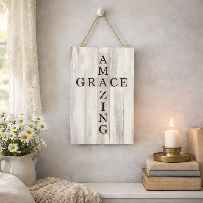 Decorative sign with 'AMAZING GRACE ZING' text hanging on a wall with a candle and flowers in the foreground.