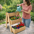 Woman watering plants in a wooden tiered garden planter outdoors.