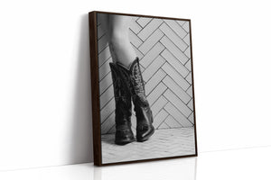 Brown wooden framed black and white photo of cowboy boots on a herringbone patterned floor.