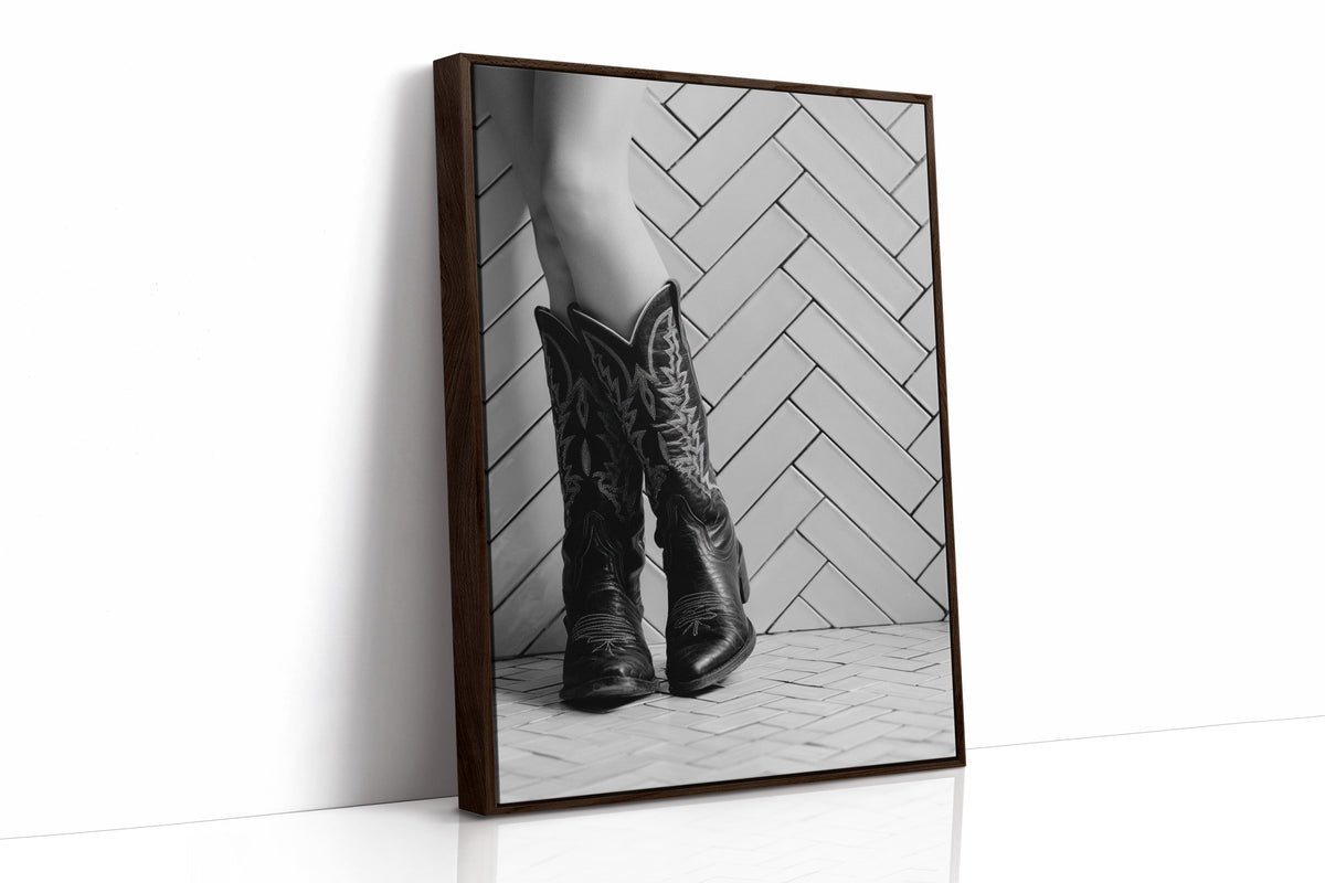Brown wooden framed black and white photo of cowboy boots on a herringbone patterned floor.
