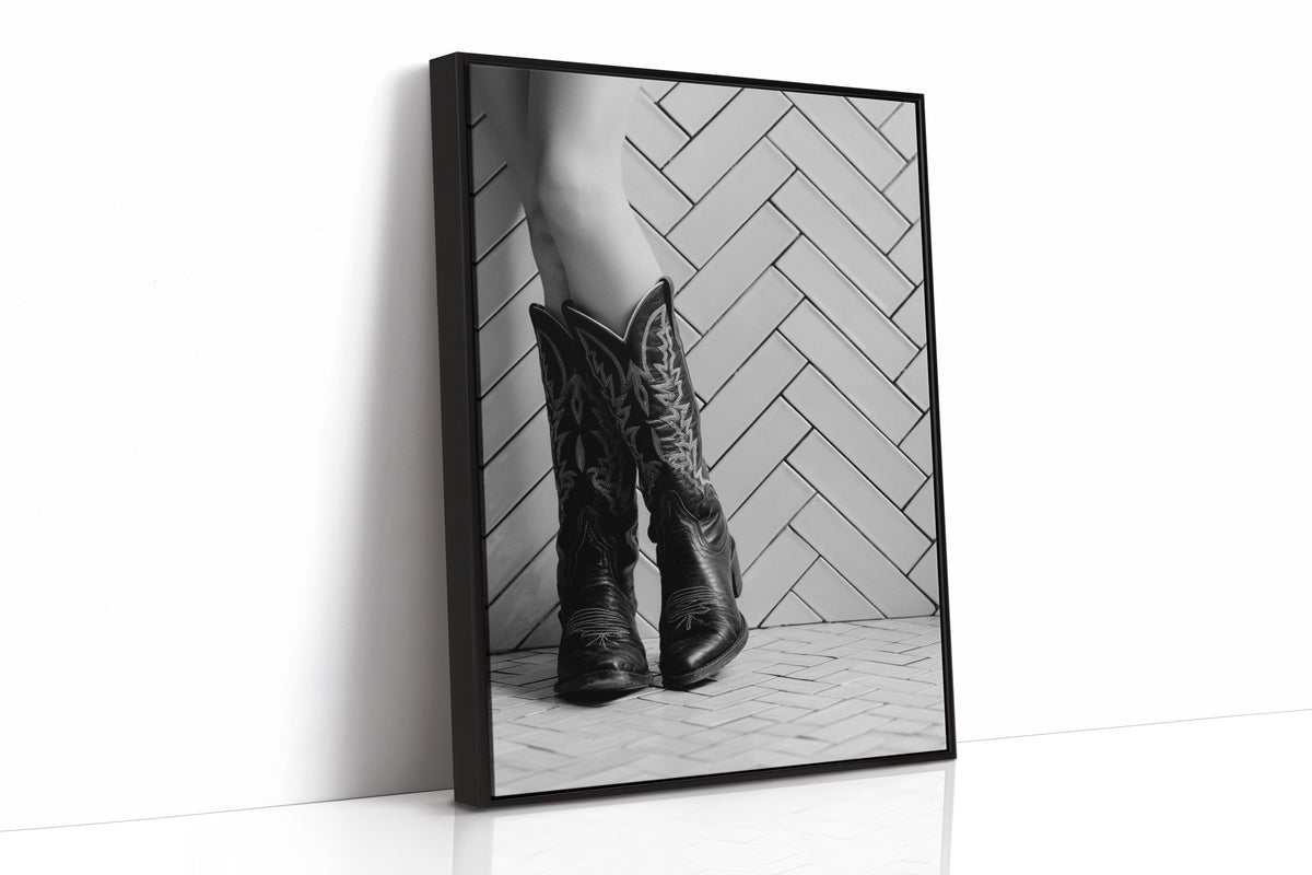 A black wooded framed black and white photographic print leaning against a white wall. The print shows a close-up of a person's legs wearing tall western-style cowboy boots in front of a herringbone-patterned wall.