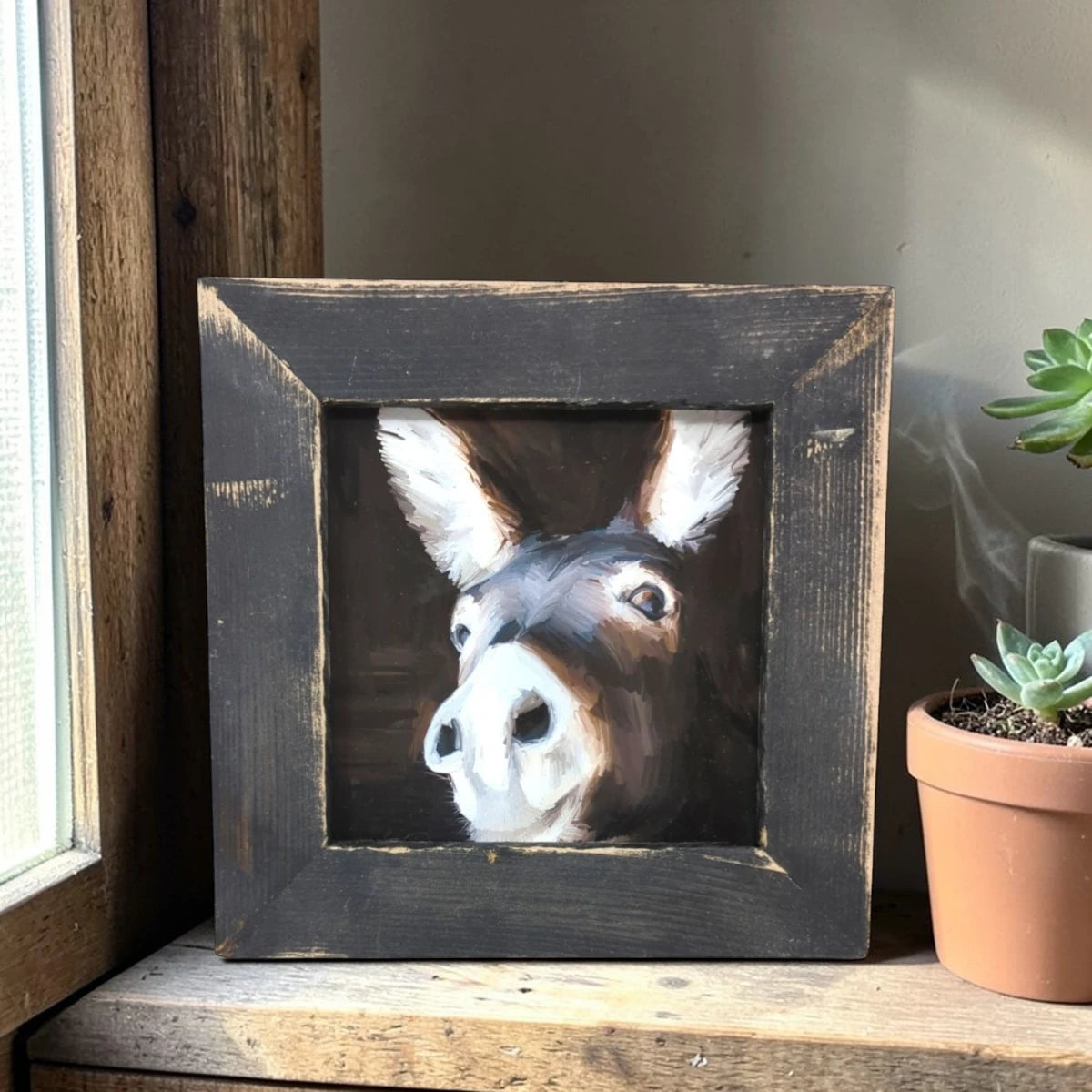Framed painting of a donkey's face on a wooden surface with a plant in the background.