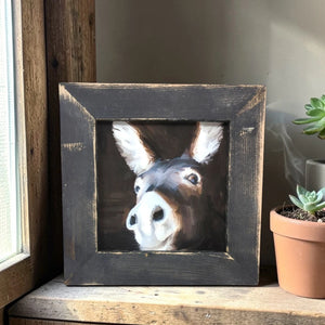 Framed painting of a donkey's face on a wooden surface with a plant in the background.