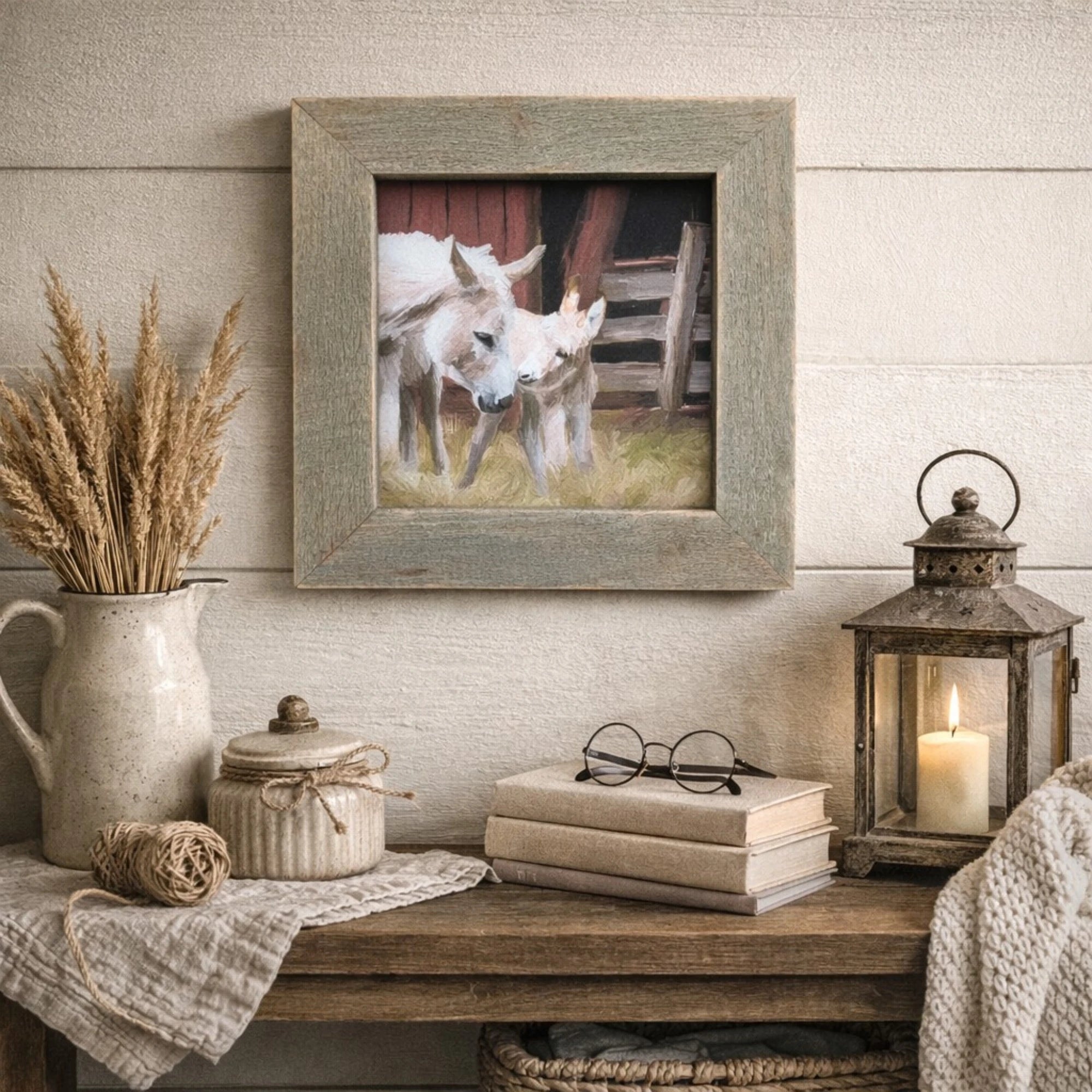 Decorative setting with a framed picture of two donkeys, a pitcher, lantern, books, and glasses on a wooden surface.