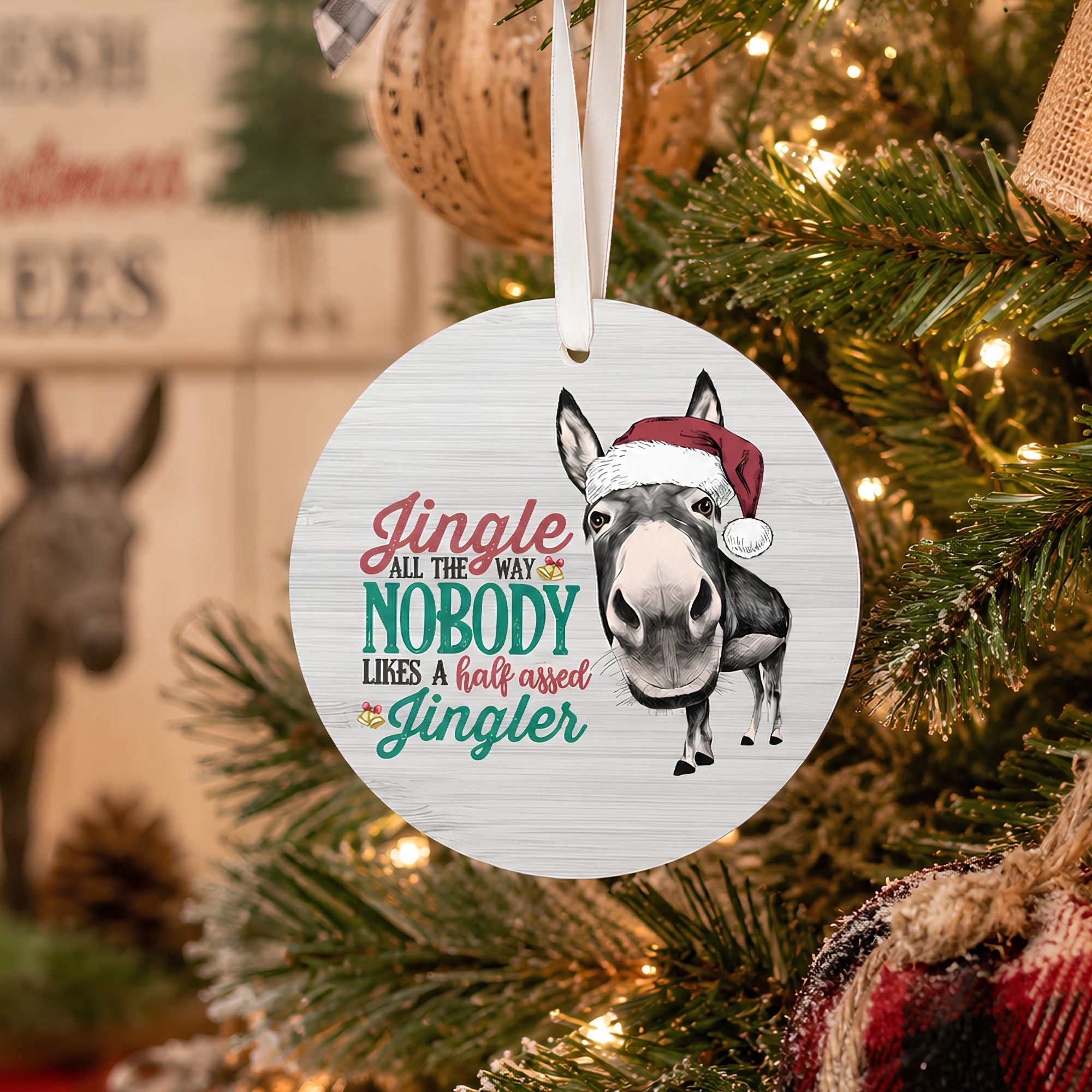 A round white Christmas ornament featuring a donkey wearing a Santa hat and the phrase 'Jingle all the way nobody likes a half assed Jingleher', hanging from a white ribbon on a decorated Christmas tree with warm lights.