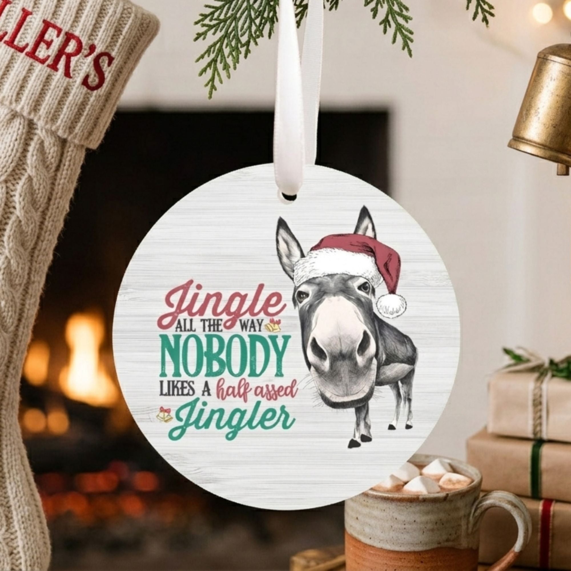 A round white Christmas ornament featuring a donkey wearing a red Santa hat and the phrase 'Jingle all the way nobody likes a half-assed jingle' in red and teal lettering. The ornament is shown hanging in a cozy holiday setting with a fireplace, a knitted stocking, wrapped gifts, and a mug of hot cocoa.