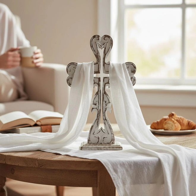 Hand-carved distressed white cross displayed as rustic farmhouse table decor with white cloth on a wooden table in a cozy living room.