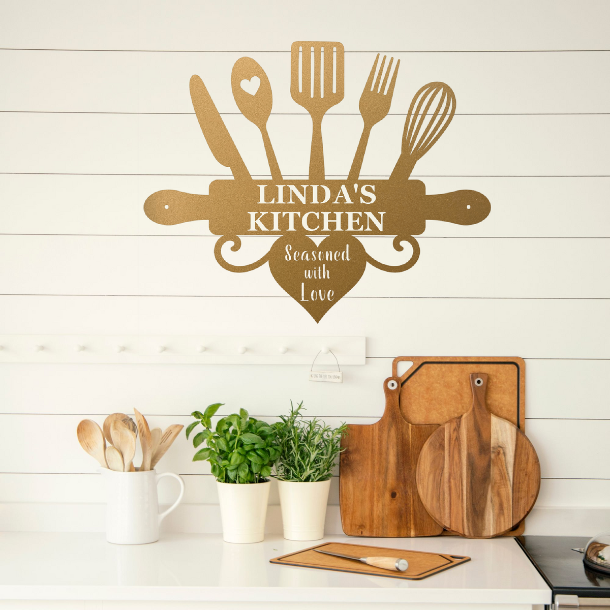 A decorative wall sign shaped like kitchen utensils and a rolling pin with the text 'LINDA'S KITCHEN' and 'Seasoned with Love' displayed above a kitchen counter with wooden cutting boards, plants, and utensils.