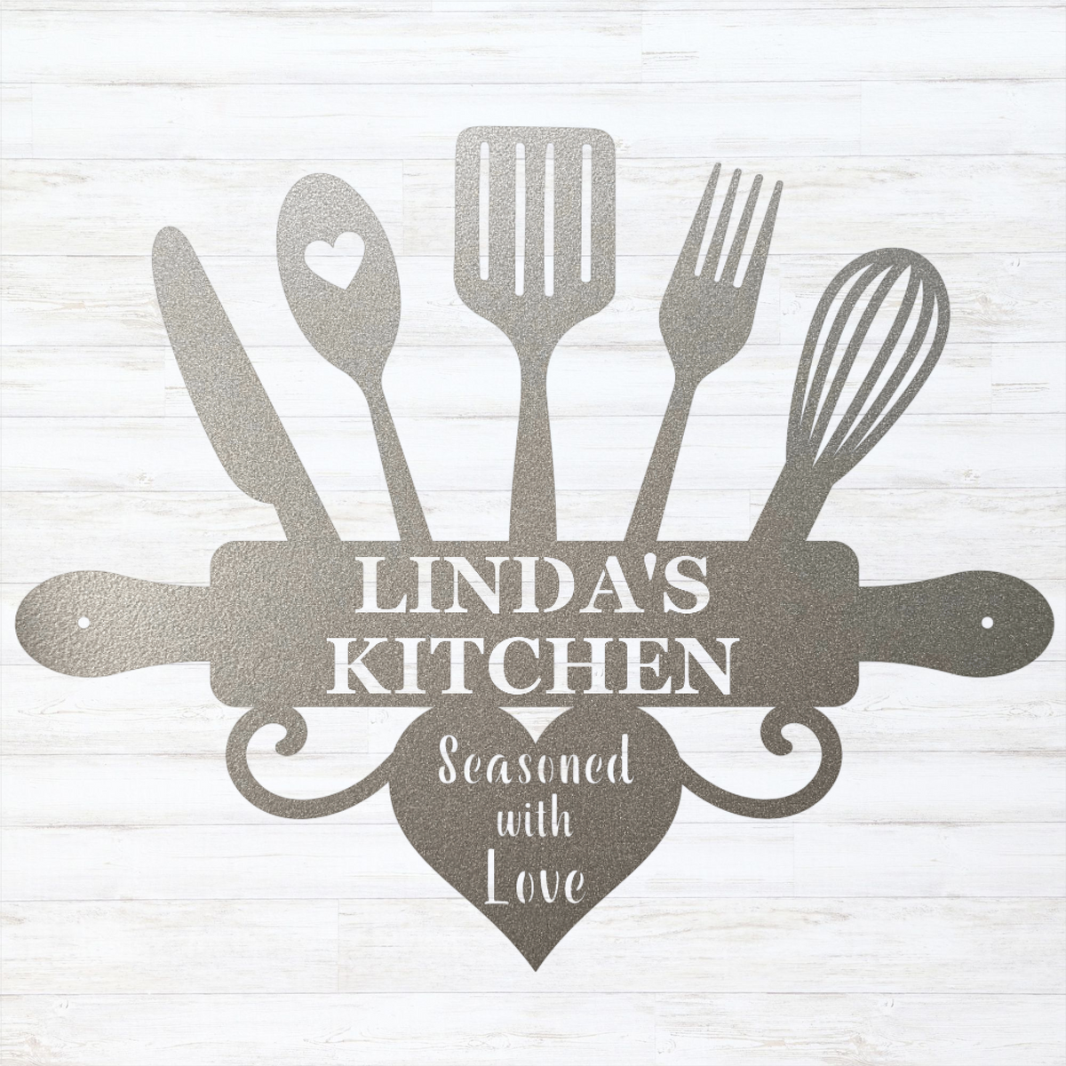 A silver personalized kitchen sign featuring silhouettes of kitchen utensils (knife, spoon with heart, spatula, fork, whisk, and rolling pin) arranged around the text 'LINDA'S KITCHEN' on a rolling pin, with a heart below reading 'Seasoned with Love'. The sign is a dark metallic gray color and is displayed against a white wood-plank background.