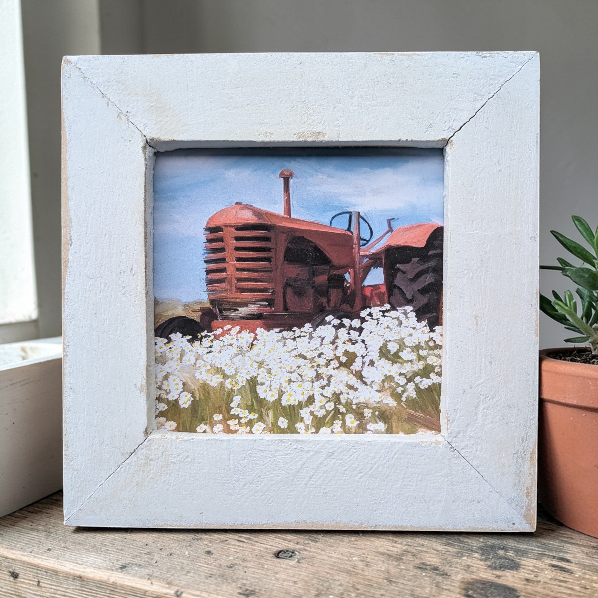 Framed picture of a red tractor in a field with white flowers on a wooden surface.