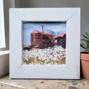 Framed picture of a red tractor in a field with white flowers on a wooden surface.