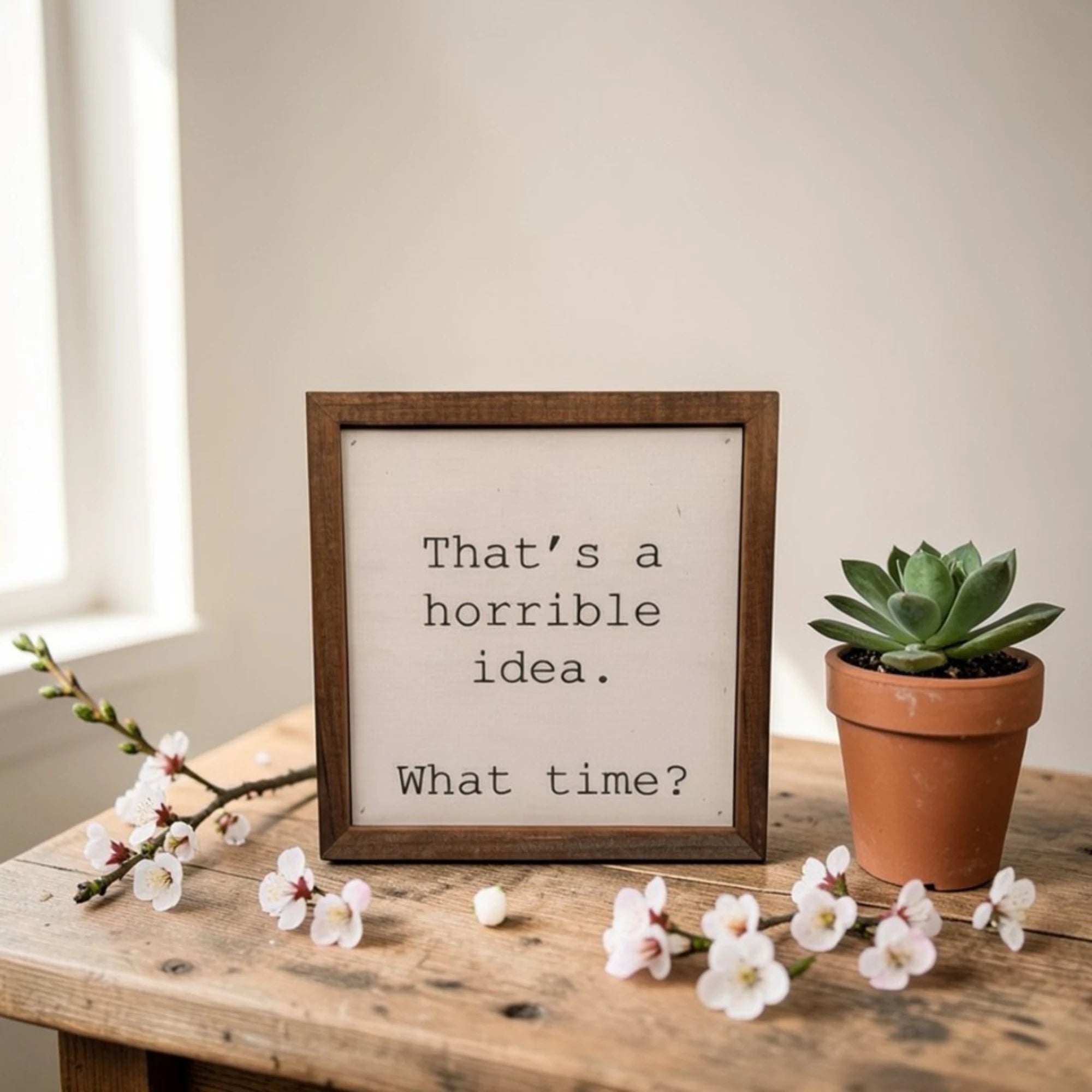 A square, wooden-framed desk sign with a white canvas face featuring the text: 'That's a horrible idea. What time?'. The sign is styled on a rustic table with flowers and a plant.