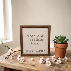 A square, wooden-framed desk sign with a white canvas face featuring the text: 'That's a horrible idea. What time?'. The sign is styled on a rustic table with flowers and a plant.