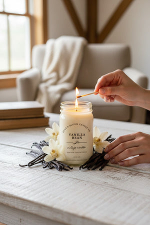 Farmhouse World Vanilla Bean Vintage Candle in a 16 oz jar on a white farmhouse-style table surrounded by vanilla beans and orchids. Creamy vanilla scent.