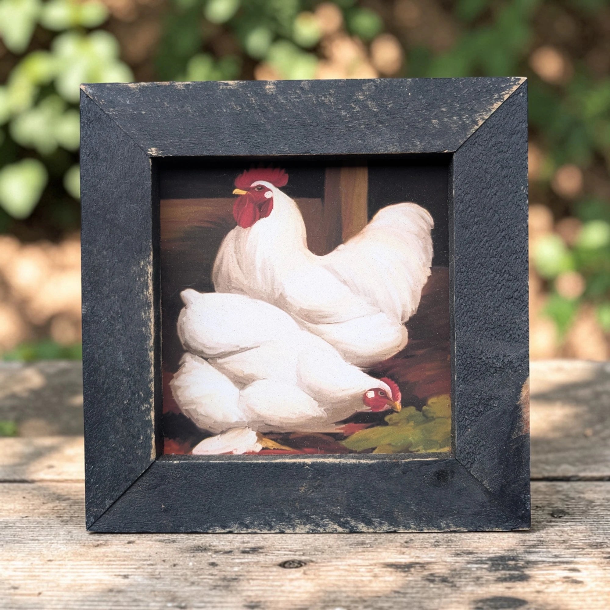 Framed artwork of two white chickens on a wooden surface with a blurred green background