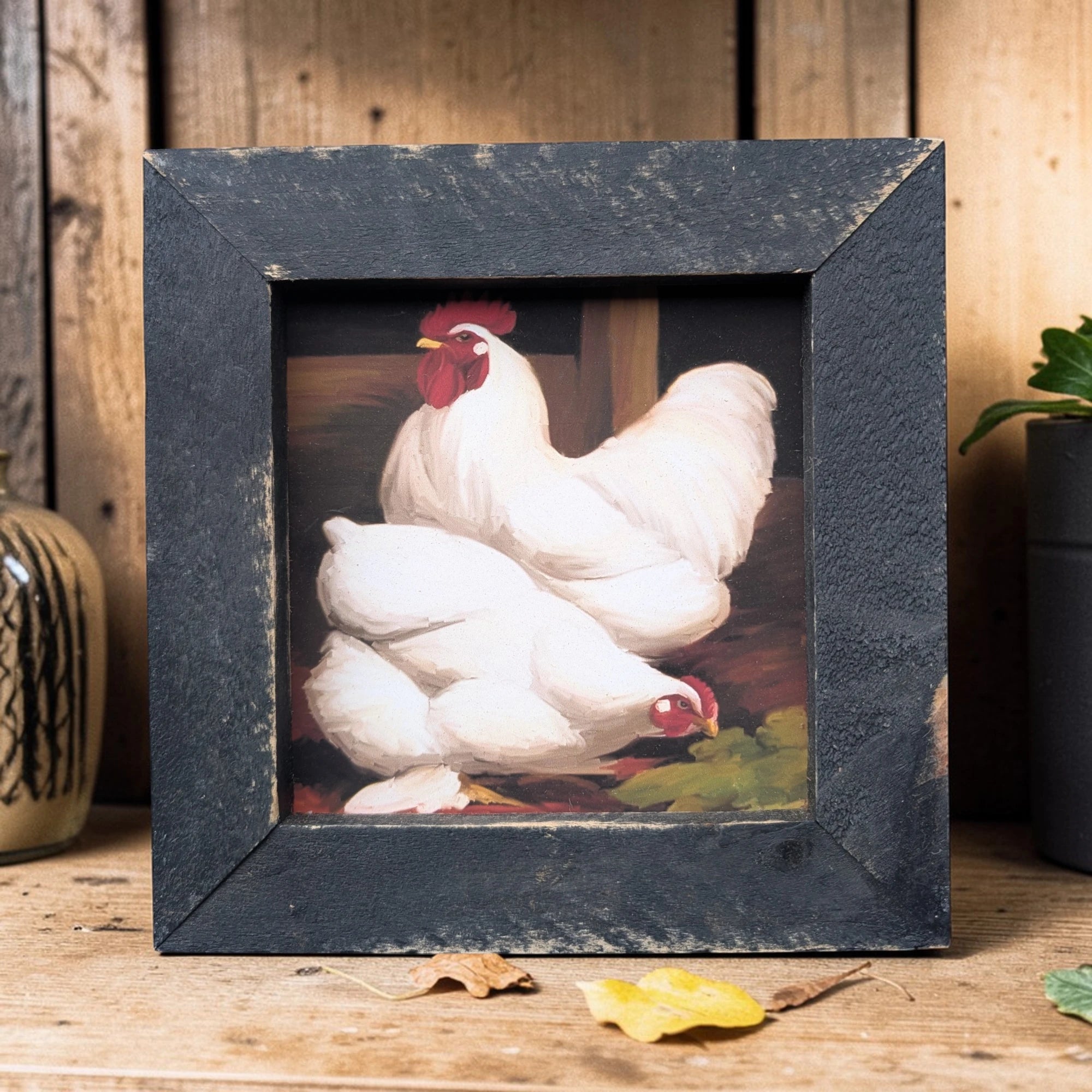 Framed artwork of two white chickens with red faces on a wooden surface.