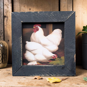 Framed artwork of two white chickens with red faces on a wooden surface.