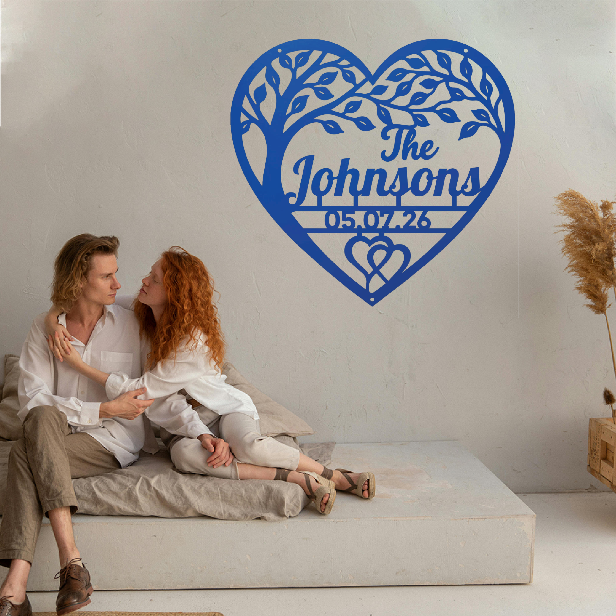 A couple sits on a low concrete bench against a white wall, with a large blue heart-shaped tree cutout wall decal behind them featuring the personalized text 'The Johnsons' and the date '05.07.