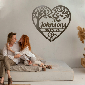 A couple sits on a low concrete bench against a light wall, with a large personalized heart-shaped tree wall art piece above them reading 'The Johnsons 05.07.