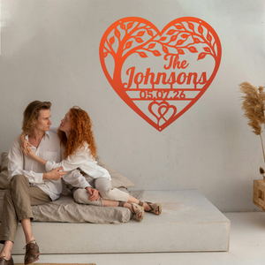 A couple sits on a low concrete bench against a light wall, with a large red heart-shaped tree silhouette wall decal behind them featuring the personalized text 'The Johnsons' and the date '05.07.