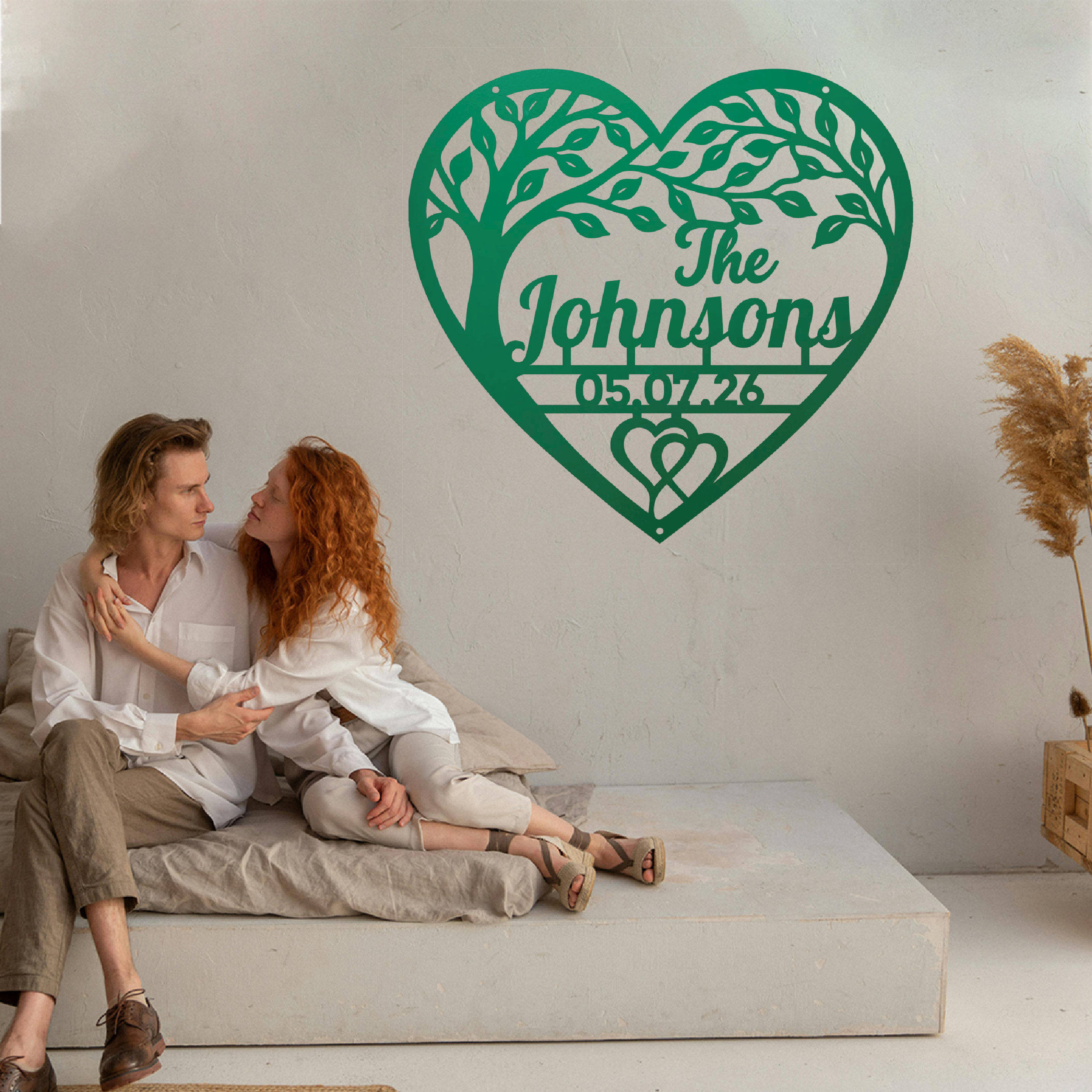 A couple sits on a low concrete bench against a white wall, with a large green heart-shaped tree silhouette and personalized text reading 'The Johnsons' and '05.07.