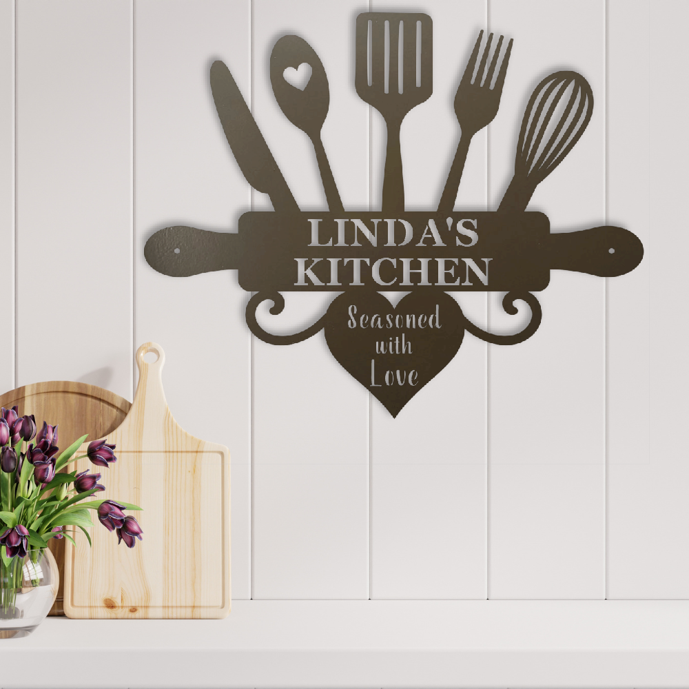 A dark brown metal kitchen wall sign shaped like a rolling pin with utensils and a heart, reading 'LINDA'S KITCHEN' and 'Seasoned with Love', mounted on a white paneled wall above a shelf with cutting boards and flowers.