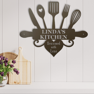 A dark brown metal kitchen wall sign shaped like a rolling pin with utensils and a heart, reading 'LINDA'S KITCHEN' and 'Seasoned with Love', mounted on a white paneled wall above a shelf with cutting boards and flowers.