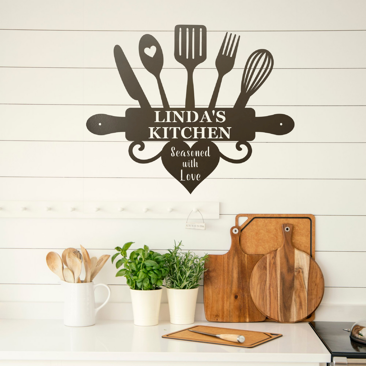 A black metal kitchen wall sign shaped like a cutting board with utensils, mounted on a white shiplap wall above a kitchen counter with cutting boards, plants, and wooden spoons.