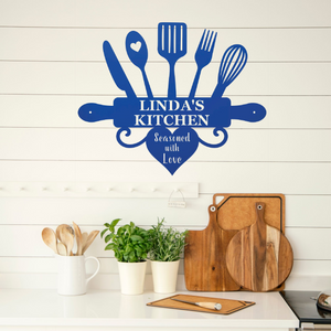 A blue kitchen utensil-shaped wall decal with the text 'LINDA'S KITCHEN' and 'Seasoned with Love' displayed on a white shiplap wall above a kitchen counter with wooden cutting boards, potted herbs, and wooden spoons.