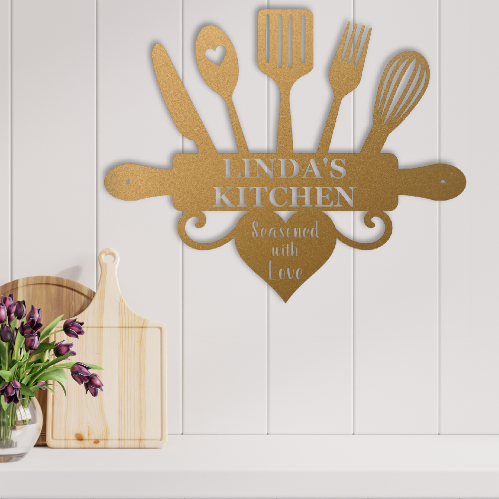 A personalized gold metal kitchen wall sign shaped like a chef's utensil set with a heart, mounted on a white paneled wall above a countertop. The sign reads 'LINDA'S KITCHEN' and 'Seasoned with Love'.