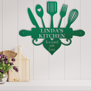 A green metal kitchen wall sign shaped like a rolling pin with utensils and a heart, reading 'LINDA'S KITCHEN' and 'Seasoned with Love,' mounted on a white paneled wall above a countertop with cutting boards and flowers.