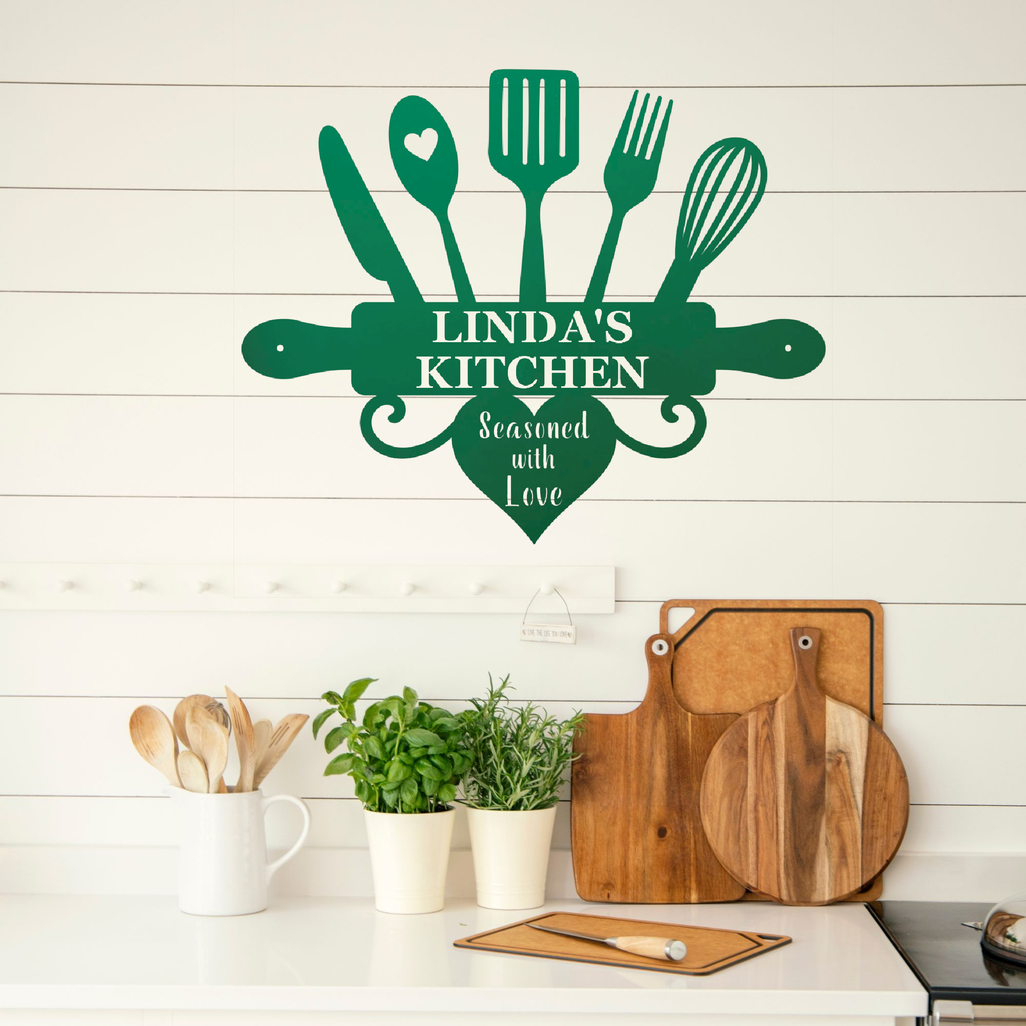 A dark green kitchen wall decal featuring kitchen utensils and the text 'LINDA'S KITCHEN' and 'Seasoned with Love' displayed on a white shiplap wall above a kitchen counter with wooden cutting boards and potted plants.
