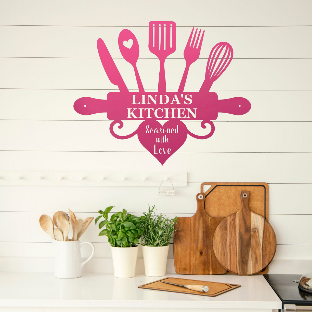 A pink kitchen-themed wall decal reading 'LINDA'S KITCHEN' and 'Seasoned with Love' is displayed on a white shiplap wall above a kitchen counter with wooden cutting boards, potted herbs, and utensils.