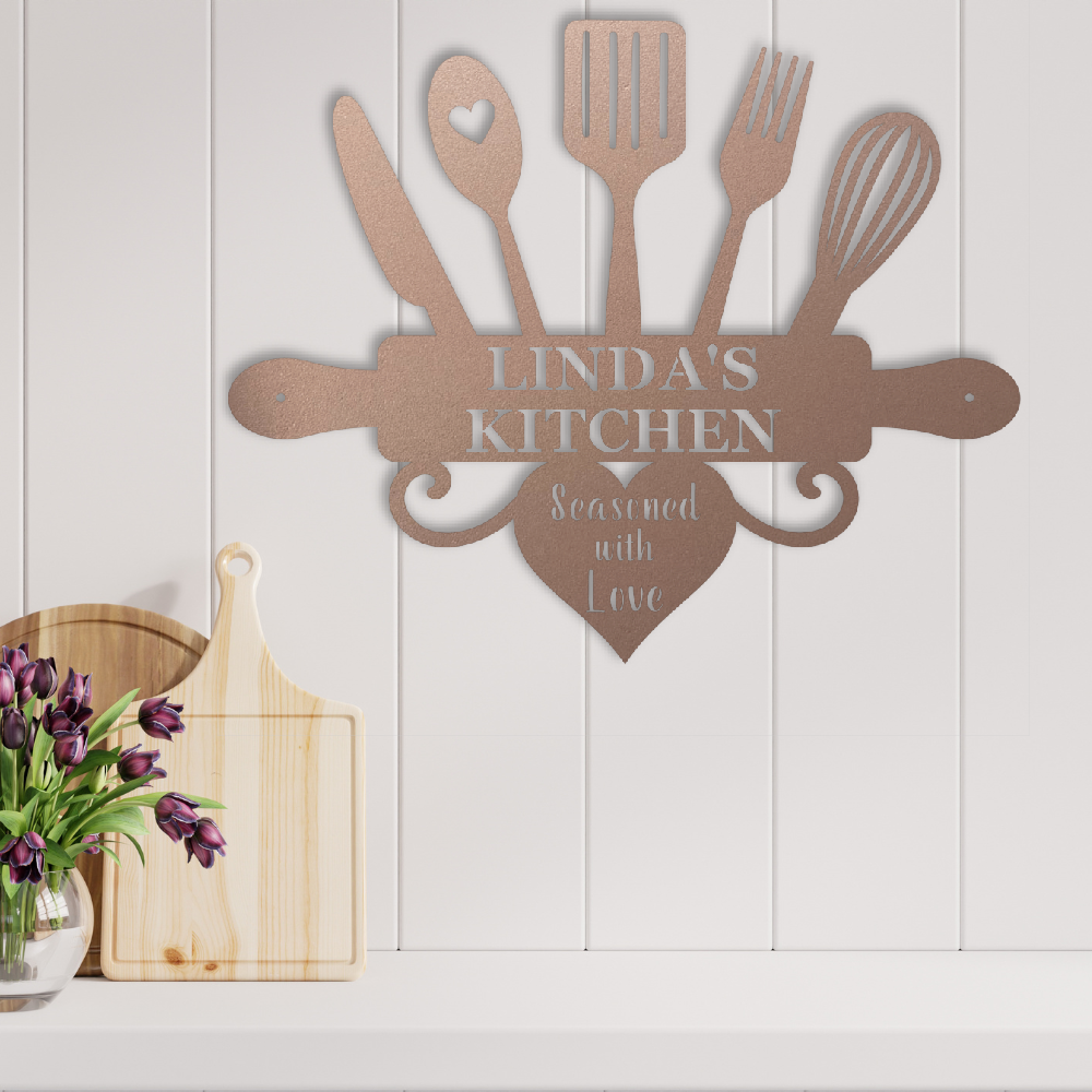 A personalized metal kitchen wall sign shaped like a rolling pin with kitchen utensils and a heart, mounted on a white paneled wall above a countertop. The sign reads 'LINDA'S KITCHEN' and 'Seasoned with Love'.