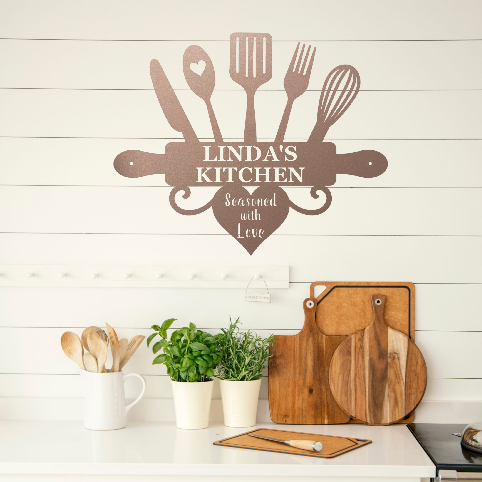A brown wall decal styled in a kitchen setting, featuring kitchen utensils and a rolling pin with the text 'LINDA'S KITCHEN' and 'Seasoned with Love'. Below the decal, a white countertop holds wooden cutting boards, a mug with wooden spoons, and potted green plants against a white shiplap wall.