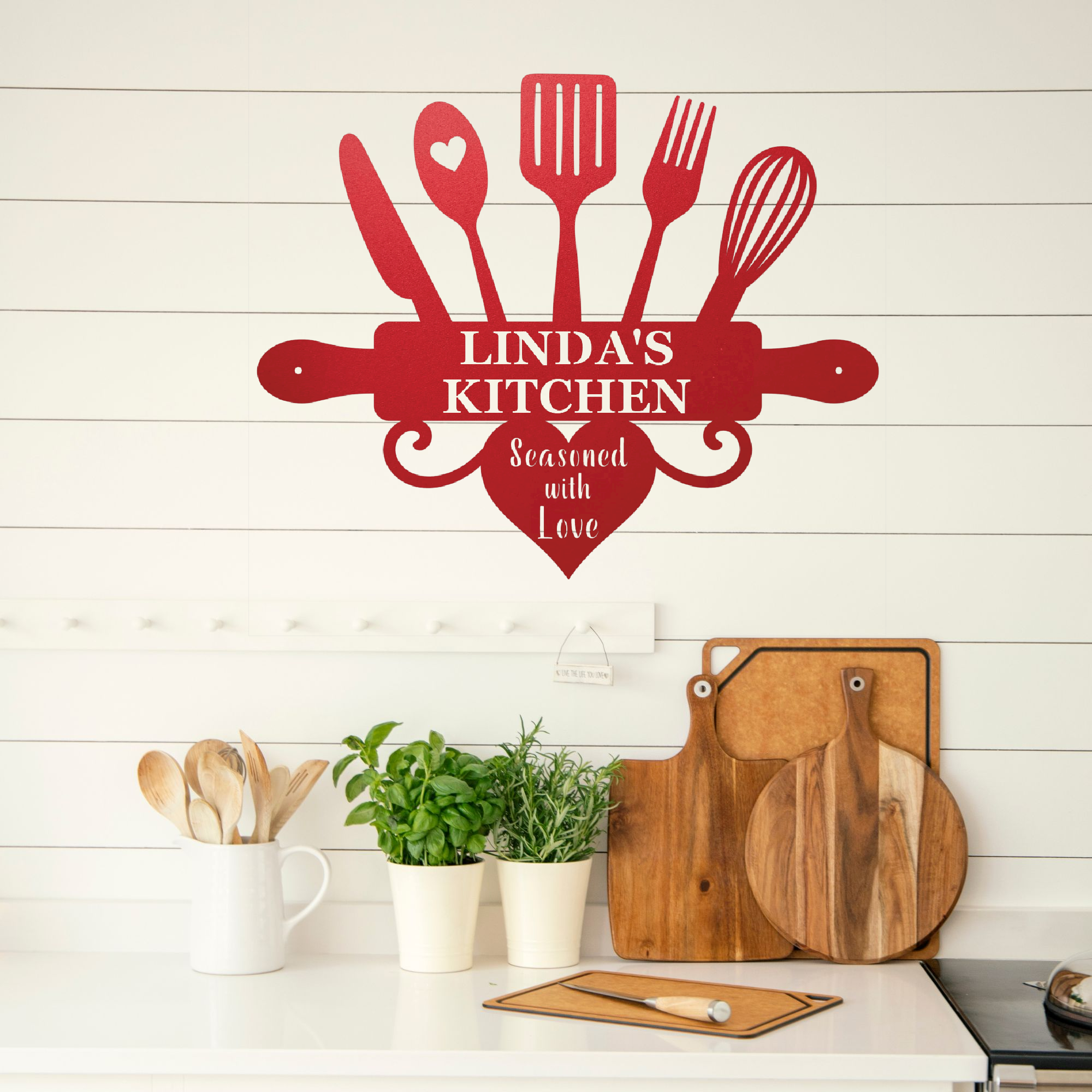A red metal kitchen wall sign shaped like a rolling pin with utensils and the words 'LINDA'S KITCHEN' and 'Seasoned with Love' displayed on a white shiplap wall above a kitchen counter with cutting boards and plants.