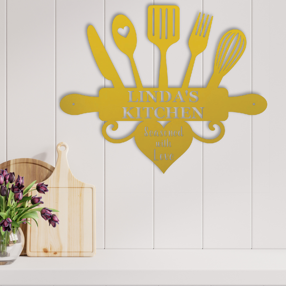 A personalized metal kitchen wall sign in mustard yellow, featuring cutout kitchen utensils and the text 'LINDA'S KITCHEN' and 'Seasoned with Love', mounted on a white paneled wall above a shelf with wooden cutting boards and flowers.