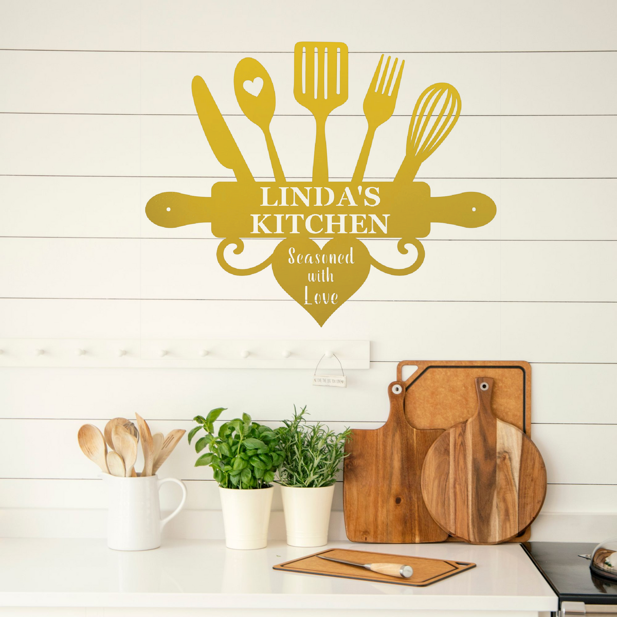 A personalized kitchen wall decal in a warm mustard yellow color, featuring kitchen utensils and the text 'LINDA'S KITCHEN' and 'Seasoned with Love'. The decal is displayed on a white shiplap wall above a kitchen counter with wooden cutting boards, potted green plants, and a white mug containing wooden spoons.